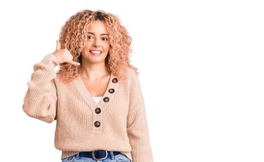 Kış süveteri giyen kıvırcık saçlı genç sarışın kadın telefonda el ve parmaklarıyla telefon hareketi yaparak telefonda konuşur gibi gülümsüyor. iletişim kavramları. 