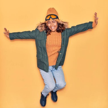 Kışlık giysiler giyen genç, güzel Latin kadın mutlu gülümsüyor. Yüzünde ve kollarında tebessümle, izole edilmiş sarı arka planda atlıyor.