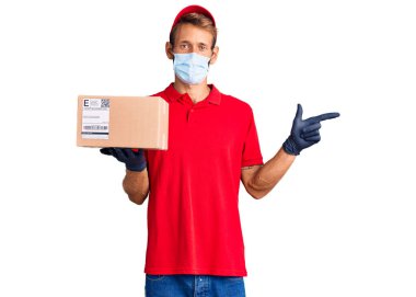 Sarışın, sakallı, elinde teslimat kutusu olan, tıbbi maske takan, gülümseyen, mutlu, eli ve parmağı yanda olan yakışıklı bir adam. 