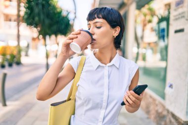 Kısa saçlı genç bayan dışarıda mutlu bir şekilde gülümsüyor ve bir fincan kahve içiyor.
