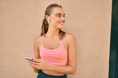 Genç sarışın sporcu şehirde akıllı telefon kullanarak egzersiz yapıyor..