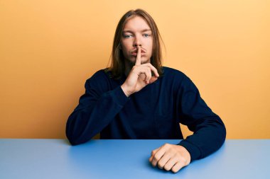 Uzun saçlı, sıradan kıyafetler giyen yakışıklı beyaz bir adam masada oturuyor ve dudakları parmaklarıyla sessiz olmak istiyor. sessizlik ve gizli konsept. 