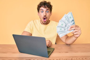 Kıvırcık saçlı, bilgisayar bilgisayarı kullanarak çalışan genç beyaz bir adam Amerikan dolarlarını elinde tutuyor. 