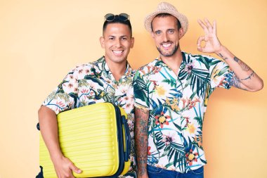 Genç eşcinsel iki adam ellerinde bavullarıyla yaz tatiline çıkıyorlar. Parmaklarıyla işaret veriyorlar, gülümseyerek mükemmel bir sembol oluşturuyorlar. 