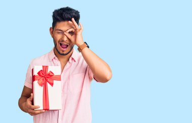 Yakışıklı Latin Amerikalı genç adam elinde hediyeyle gülümsüyor mutlu bir şekilde el pençe divan duruyor. 