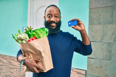 Sakallı Afrikalı Amerikalı bir adam elinde süpermarketten alınmış kağıt torbalarla kredi kartı gösteriyor.