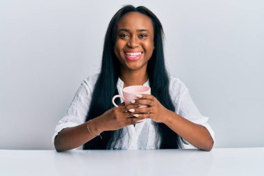 Genç Afrikalı bir kadın kahve içiyor mutlu ve serin bir gülümsemeyle gülümsüyor. diş gösterme. 