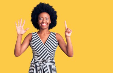 Genç Afro-Amerikalı kadın gündelik kıyafetler giyip altı numaralı parmaklarıyla işaret ederken kendinden emin ve mutlu bir şekilde gülümsüyor.. 