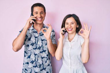 Akıllı telefondan konuşan güzel bir çift parmaklarıyla imza atıyor, gülümsüyor ve mükemmel bir sembol oluşturuyor. 