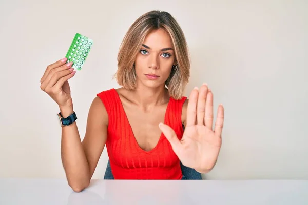 Beautiful caucasian woman holding birth control pills with open hand doing stop sign with serious and confident expression, defense gesture