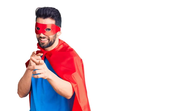 Young handsome man with beard wearing super hero costume pointing fingers to camera with happy and funny face. good energy and vibes.