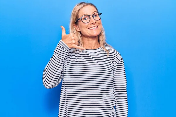 Middle age caucasian blonde woman wearing casual striped sweater and glasses smiling doing phone gesture with hand and fingers like talking on the telephone. communicating concepts.