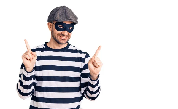 強盗マスクを身に着けている若いハンサムな男は 異なる方向に指で自信を持って指して笑っている 広告のコピースペース — ストック写真