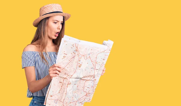 Young beautiful girl wearing hat holding city map scared and amazed with open mouth for surprise, disbelief face
