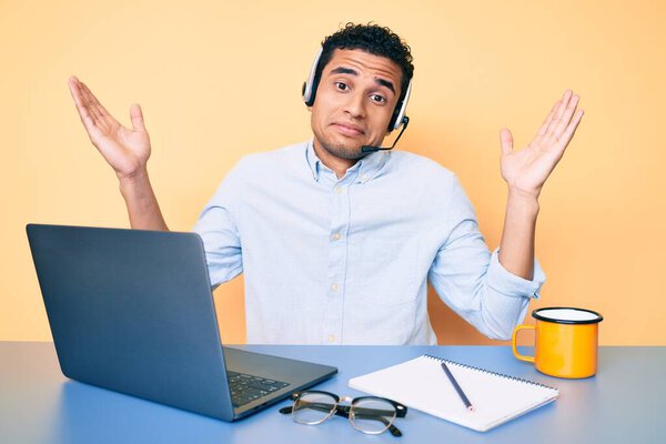 Молодой красивый латиноамериканец, работающий в офисе в наушниках оператора, невежественный и смущенный с открытыми объятиями, без понятия и сомнительным лицом. 