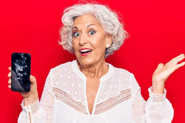 Senior grey-haired woman holding broken smartphone showing cracked screen celebrating achievement with happy smile and winner expression with raised hand