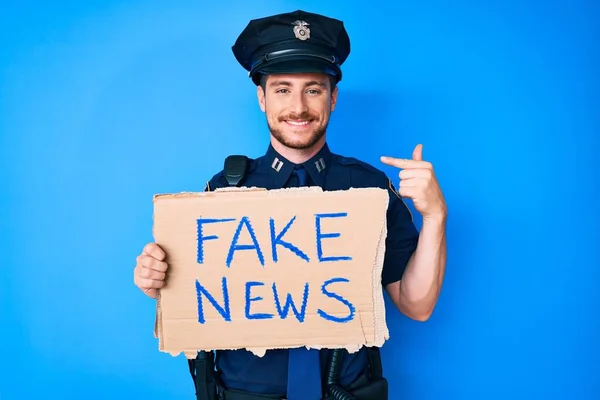 Young caucasian man wearing police uniform holding fake news banner pointing finger to one self smiling happy and proud