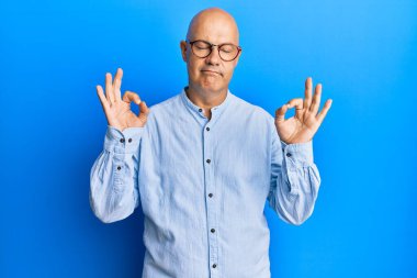 Orta yaşlı kel bir adam günlük kıyafetler ve gözlük takıyor rahatlıyor ve gözleri kapalı gülümsüyor parmaklarıyla meditasyon hareketi yapıyor. yoga konsepti. 