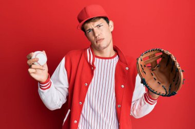Beyzbol üniforması giyen yakışıklı beyaz adam Golve 'u ve topu tutuyor ve kafası karışık bir ifade takınıyor. şüphe kavramı. 