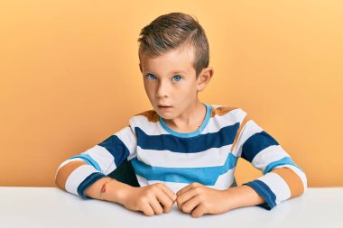 Adorable caucasian kid wearing casual clothes sitting on the table in shock face, looking skeptical and sarcastic, surprised with open mouth  clipart