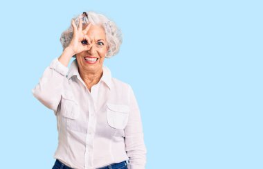 Yaşlı, gri saçlı, günlük kıyafetler giyen, el hareketleriyle gülümseyen, mutlu yüzlü parmaklarının arasından bakan bir kadın.. 