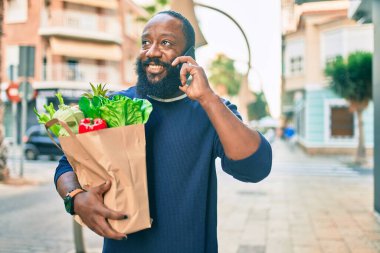 Elinde süpermarketten alınmış kağıt torba sakallı Afrikalı Amerikalı adam telefonda konuşuyor.