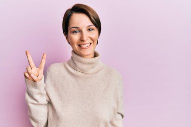 Kısa saçlı esmer kadın, günlük kışlık süveter giyiyor ve iki numaralı parmaklarıyla işaret ediyor. Kendinden emin ve mutlu bir şekilde gülümsüyor.. 