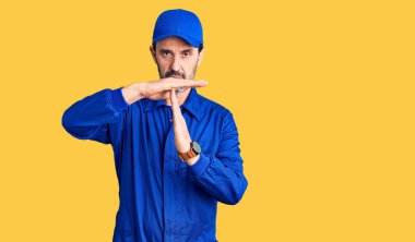 Orta yaşlı, tamirci üniforması giyen yakışıklı bir adam. El kol hareketi yapıyor, hayal kırıklığına uğramış ve ciddi bir yüz ifadesi var. 