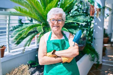 Gri saçlı, eldivenli ve bahçıvan önlüklü yaşlı kadın evdeki bitkileri bahçeliyor.