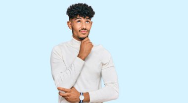 Genç Arap adam, sıradan bir kazak giyiyor, elleri çenesinde soru soruyor, düşünceli bir yüz ifadesi var. Gülümseyen ve düşünceli bir yüz. şüphe kavramı. 