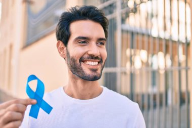 Genç İspanyol gülümseyen adam şehirde mavi kurdele tutarken mutlu..