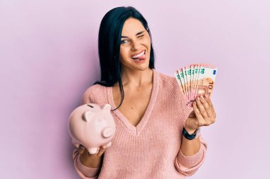 Young caucasian woman holding euro banknotes and piggy bank winking looking at the camera with sexy expression, cheerful and happy face. 