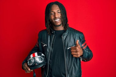 Saçları örgülü, elinde motosiklet kaskı olan, mutlu ve pozitif gülümseyen, başparmağıyla mükemmel ve onay işareti yapan Afro-Amerikan bir adam. 