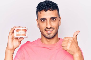 Takma dişleri olan genç İspanyol adam mutlu ve pozitif gülümsüyor, başparmağıyla mükemmel ve onay işareti yapıyor. 