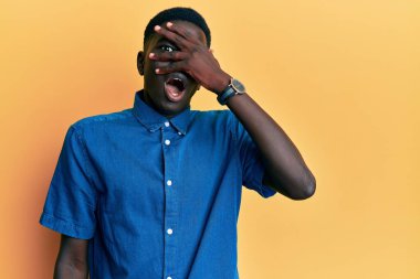 Genç Afro-Amerikan adam günlük giysiler giyip şok içinde yüzünü ve gözlerini eliyle kapatıyor, parmaklarından korkuyla bakıyor. 