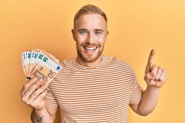 Elinde 50 avroluk banknotlar tutan genç beyaz adam bir fikir veya soru işareti ile gülümsüyor. 