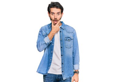 Gündelik kıyafetler giyen genç İspanyol adam, elleri çenesinde, inanamama, şaşkınlık ve şaşkınlık dolu bir ifade takınıyor. 