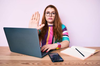 Ofiste laptopuyla çalışan genç kızıl saçlı kadın ciddi ve kendinden emin bir ifadeyle dur işareti yapıyor, savunma jesti. 