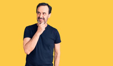 Orta yaşlı yakışıklı bir adam, elleri çenesinde gündelik tişörtler giyip soru soruyor, düşünceli bir yüz ifadesi var. Düşünceli bir yüzle gülümsüyor. şüphe kavramı. 