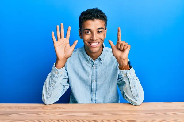 若いですハンサムなアフリカ系アメリカ人男性を着てカジュアルな服を着てテーブルの上に座って指の番号7を示すと指摘しながら 自信と幸せ — ストック写真