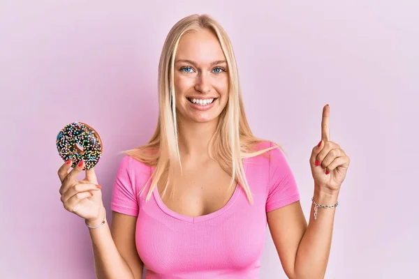 Jovem Loira Segurando Donut Sorrindo Com Uma Ideia Pergunta Apontando — Fotografia de Stock