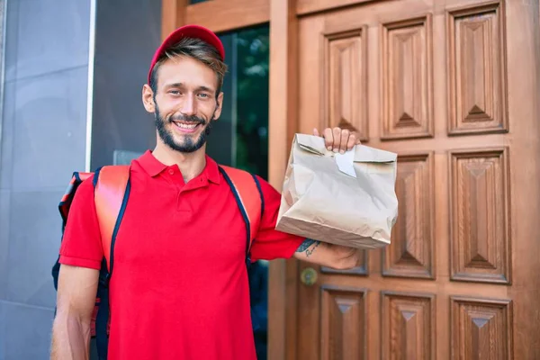 白种人送货员身穿红色制服 背着外卖背包 心情愉快地微笑着 拿着纸袋 — 图库照片