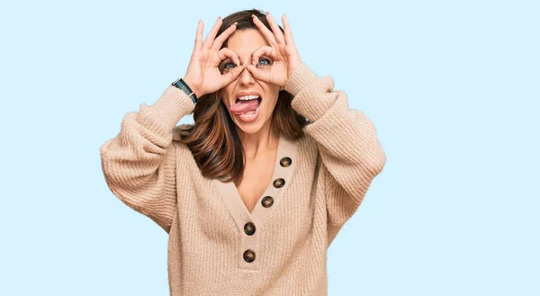 若いブルネットの女性は 双眼鏡が舌を突き出すようなOkジェスチャーをするカジュアルな冬のセーターを着て 目は指を通して見ている クレイジーな表現 — ストック写真
