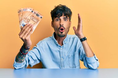 Young hispanic man holding 50 euro banknotes sitting on the table scared and amazed with open mouth for surprise, disbelief face 