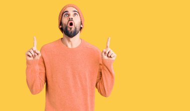 Sakallı, sıradan bir kazak ve yün şapka giyen genç yakışıklı adam şaşırmış ve şaşırmış bir şekilde yukarı bakıyor ve parmaklarıyla işaret ediyor ve kollarını kaldırıyor.. 