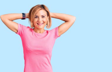 Spor giyinen genç sarışın kadın rahatlıyor ve esniyor, kolları ve elleri başının arkasında ve boynunda mutlu bir şekilde gülümsüyor. 