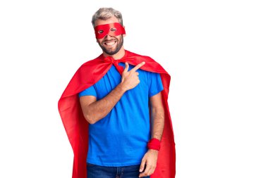 Sarışın, süper kahraman kıyafeti giyen, neşeli, yüzünde mutlu ve doğal bir ifadeyle, elleri ve parmaklarıyla yüzünü gösteren bir gülümseme olan genç bir adam. 
