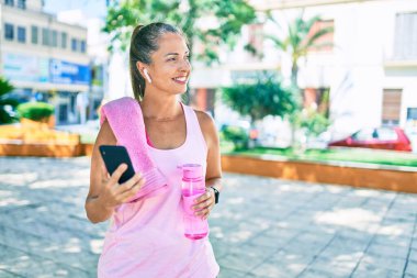 Parkta akıllı telefon kullanarak gülümseyen orta yaşlı sporcu kadın.