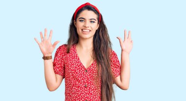 Yazlık elbise giyen esmer kız on numarayı gösteriyor ve parmaklarıyla işaret ediyor. Kendinden emin ve mutlu bir şekilde gülümsüyor.. 