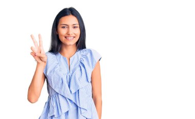 Gündelik kıyafetler giymiş, gülümseyen, parmaklarını zafer işareti yaparken gösteren genç ve güzel bir Latin kız. İki numara.. 